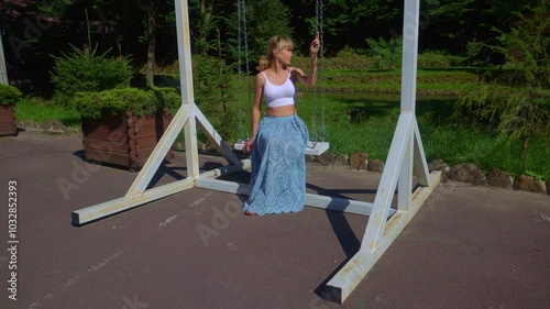 A woman swings on a wooden swing. A woman sits on a swing in the park and squints in the sun.