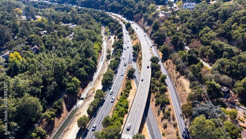 Aerial view of moderate traffic on multi-lane highway 17 surrounded by dense trees and residential houses. Power lines stretch along the road, with Los Gatos Creek flowing nearby