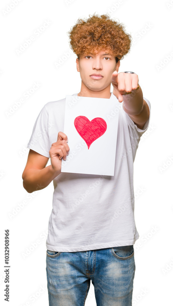 Young handsome man holding paper with red heart pointing with finger to the camera and to you, hand sign, positive and confident gesture from the front Young handsome man holding paper with red heart pointing with finger to the camera and to you, hand sign, positive and confident gesture from the front