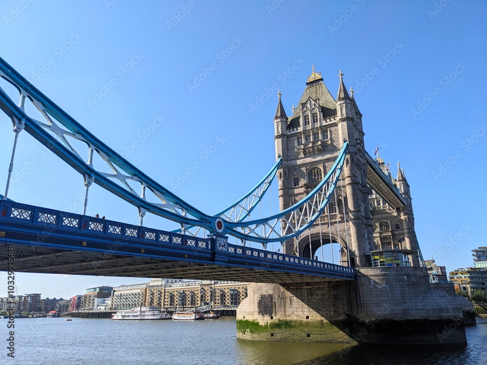 Obraz premium London, UK - 2023.07.02: Side view of Tower Bridge with Queen Elizabeth II Silver Jubilee repainting of blue and white over River Thames under a clear blue sky with sunlight coming from the left