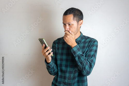 Young men wearing green flannel covering his mouth while looking smartphone on isolated white background