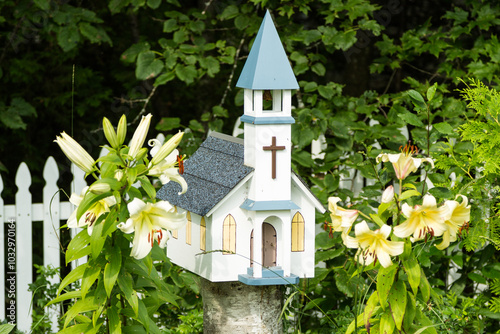 A miniature model church birdhouse on a birch tree stump, with lilies on each side and a white picket fence in the background. photo