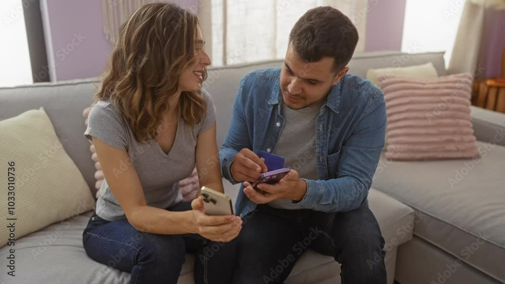 Couple using smartphones and credit card while sitting on a couch in a cozy living room, smiling and enjoying their time together at home Couple using smartphones and credit card while sitting on a couch in a cozy living room, smiling and enjoying their time together at home