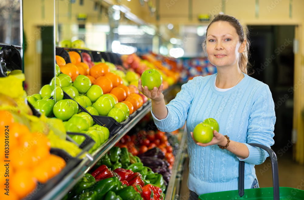 Obraz premium Pleased middle-aged woman buyer purchasing fresh apples at the counter in large vegetable shop