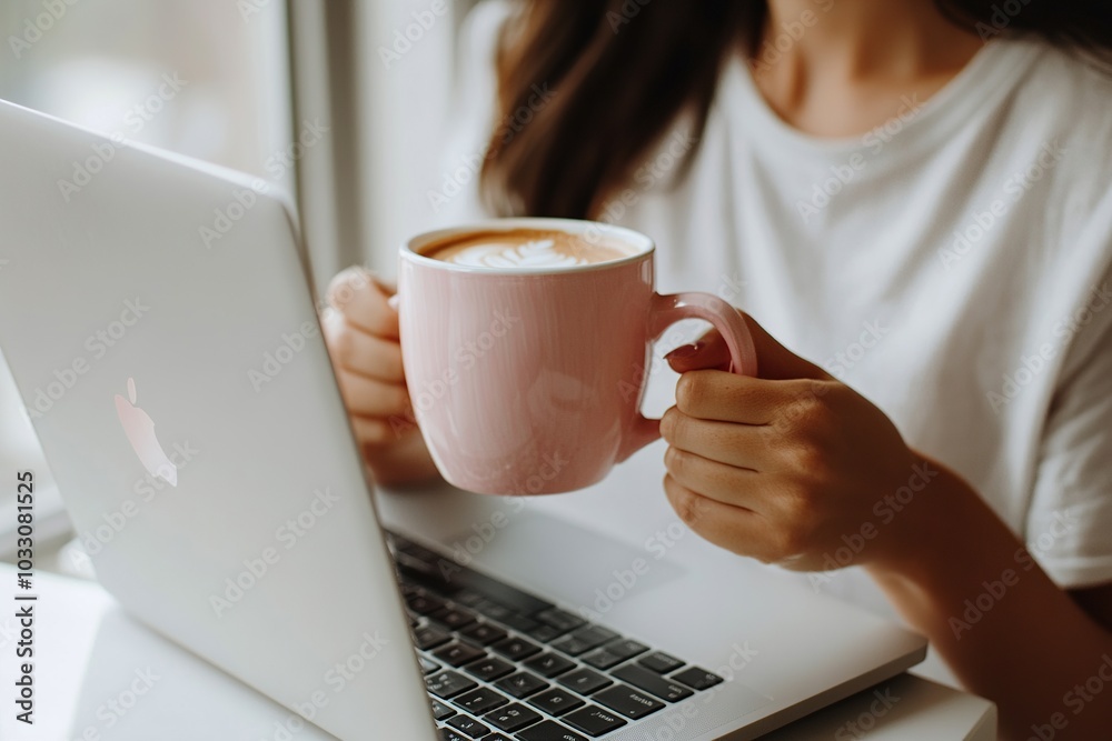 A woman sits at her desk with an open laptop, holding and drinking coffee from a pink cup on top of it A woman sits at her desk with an open laptop, holding and drinking coffee from a pink cup on top of it