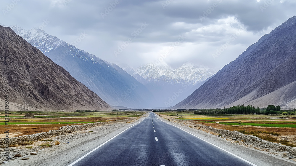 Naklejka premium A straight road leading into the mountains cuts through a wide valley between majestic snow-capped mountain ranges. In the distance, a cloudy sky looms on the horizon