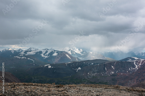 Dramatic top view from high stony pass to alpine valley against large mountain range with snow-capped peaks in gray rainy low clouds. Beautiful big snowy mountain tops in rain under grey cloudy sky.