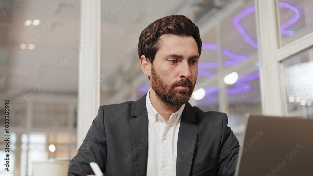 A businessman sits in a cafe in front of his laptop, writing on a notepad. He is wearing a suit, shirt and has a beard. The cafe is modern with glass partitions and purple neon lighting. A businessman sits in a cafe in front of his laptop, writing on a notepad. He is wearing a suit, shirt and has a beard. The cafe is modern with glass partitions and purple neon lighting.