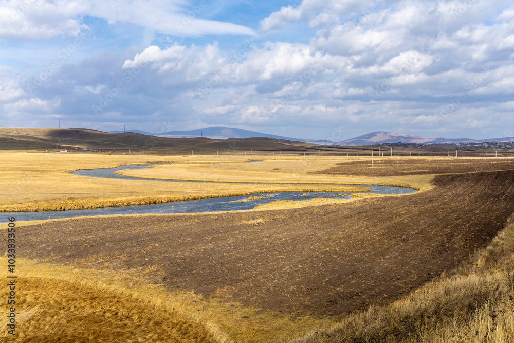 Plowed field near a winding river flowing through fields of dry yellow grass. Hills and a bright sky with clouds in the background Plowed field near a winding river flowing through fields of dry yellow grass. Hills and a bright sky with clouds in the background