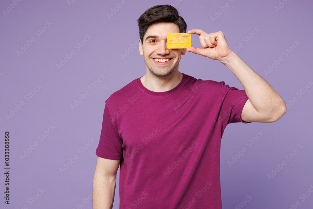 Young Caucasian man he wear violet t-shirt casual clothes hold in hand cover eye with mock up of credit bank card isolated on plain pastel light purple background studio portrait. Lifestyle concept. Young Caucasian man he wear violet t-shirt casual clothes hold in hand cover eye with mock up of credit bank card isolated on plain pastel light purple background studio portrait. Lifestyle concept.