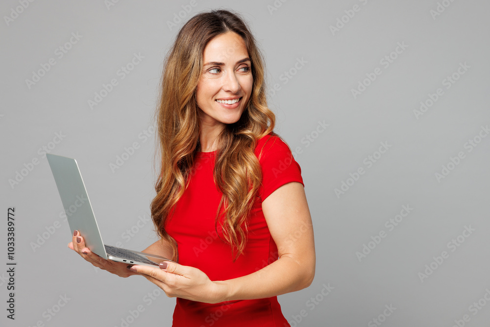 Side view young happy IT woman she wear red t-shirt casual clothes hold use work on laptop pc computer look aside on area mock up isolated on plain grey background studio portrait. Lifestyle concept. Side view young happy IT woman she wear red t-shirt casual clothes hold use work on laptop pc computer look aside on area mock up isolated on plain grey background studio portrait. Lifestyle concept.