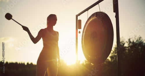 Standing in the Warm Evening Light, Serene Woman Hits a Metal Gong, Letting Its Soothing Resonance Deepen Her Relaxation. Vibrations Blend with Peaceful Outdoors, Creating a Meditative Atmosphere