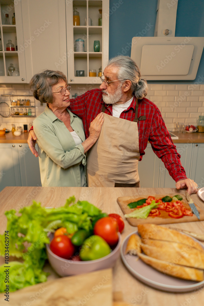 in bright kitchen an elderly woman in a light green shirt with her husband in an apron are standing in the kitchen hugging, smiling, looking at the camera in bright kitchen an elderly woman in a light green shirt with her husband in an apron are standing in the kitchen hugging, smiling, looking at the camera