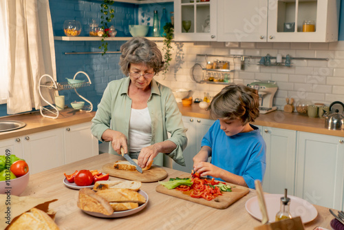 in a bright kitchen an elderly woman in a light green shirt teaches her grandson in blue T-shirt to cut vegetables for a salad