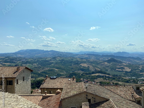 View from above on the roofs of the ancient houses of San Marino, above which the boundless mountain horizon stretches against the background of the sunny blue sky.