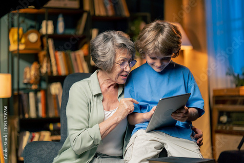 a warm light of living room with library big blue armchair elderly woman sitting with grandson watching news from tablet modern grandmother