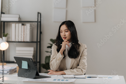 Focused Businesswoman: A thoughtful young Asian businesswoman sits at her desk, reviewing documents on her tablet, showcasing professionalism and concentration in a modern office setting. 