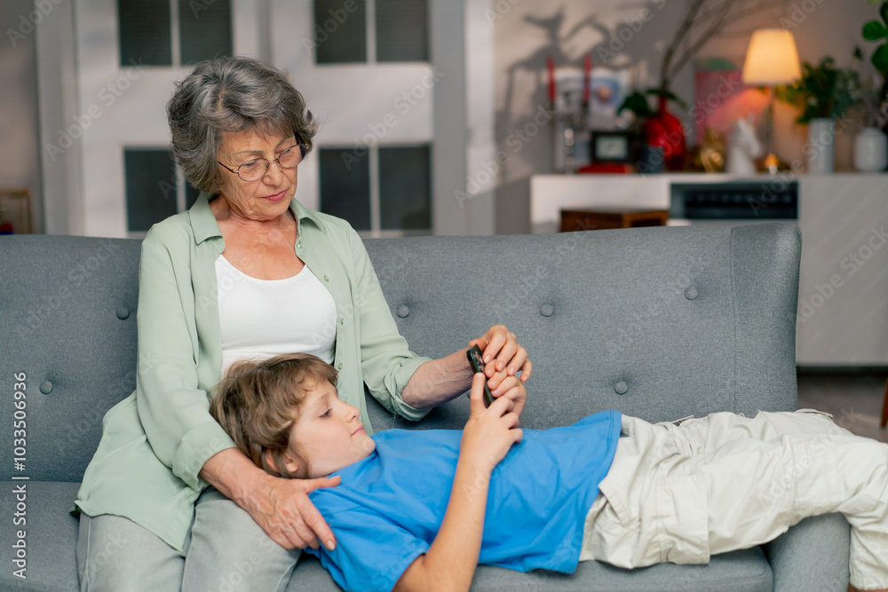 Obraz premium bright living room with sofa grandmother sitting on sofa grandson lying and watching cartoons on tablet happy family