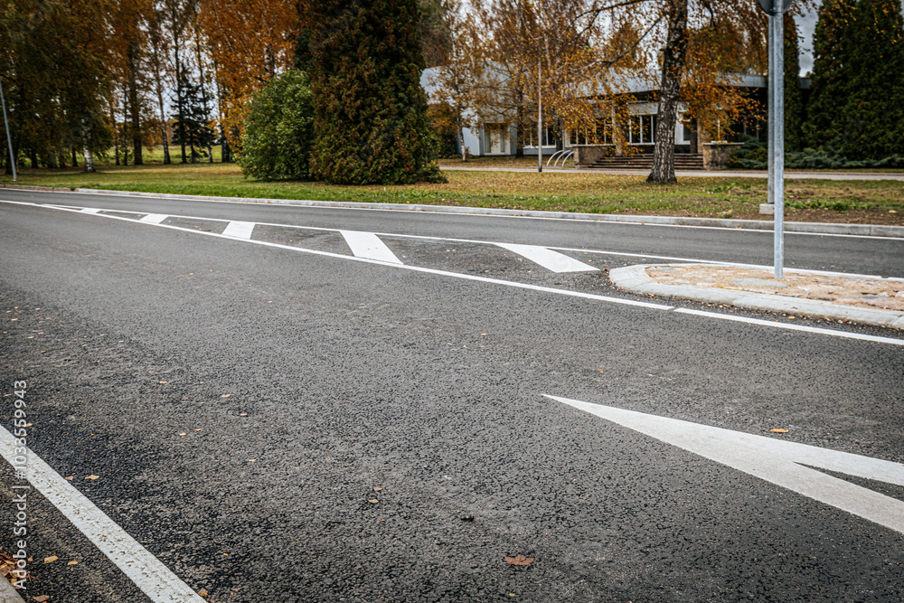 Obraz premium A quiet, empty asphalt road with a white directional arrow and painted lane markings. The scene shows fall foliage and trees in the background, with a nearby building partially visible.