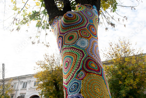 A tree wrapped in colorful crochet yarn art, showcasing vibrant patterns against a backdrop of an urban park. A unique blend of nature and creative street art in autumn