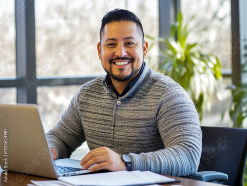 A happy Hispanic man in an office, confidently sitting at a desk with a laptop and papers, smiling at the camera.