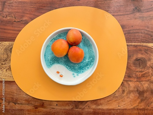 Top view of three fresh peaches in a ceramic blue bowl, placed on an orange placemat over a wooden table, creating a vibrant and colorful contrast.