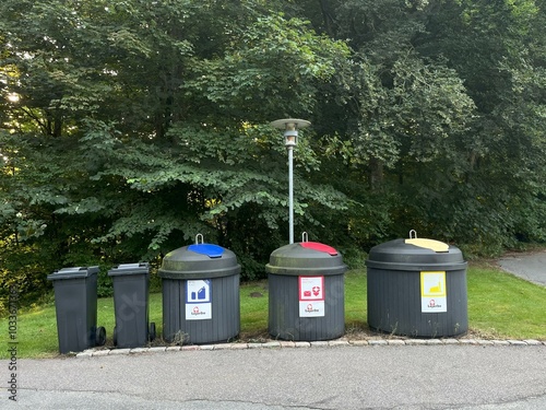 Outdoor recycling station with three large bins for sorting glass, plastic, and metal waste, alongside standard trash bins, set against a backdrop of lush green trees in a park.