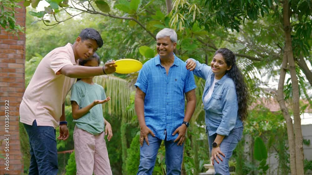 Indian Asian family of four spending quality time outdoors in their home lawn, parents in their forties watching as teenage son teaches his little sister how to throw a frisbee or flying disc Indian Asian family of four spending quality time outdoors in their home lawn, parents in their forties watching as teenage son teaches his little sister how to throw a frisbee or flying disc