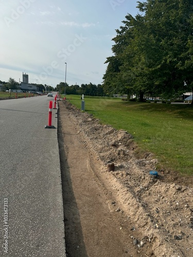 Roadside construction with traffic cones and freshly dug trench along a tree-lined street, under clear skies. Vehicles visible in the distance near a building.