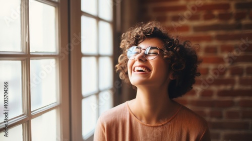 A young woman with short, curly hair and glasses beams with joy near a sunlit window, radiating happiness and vitality.