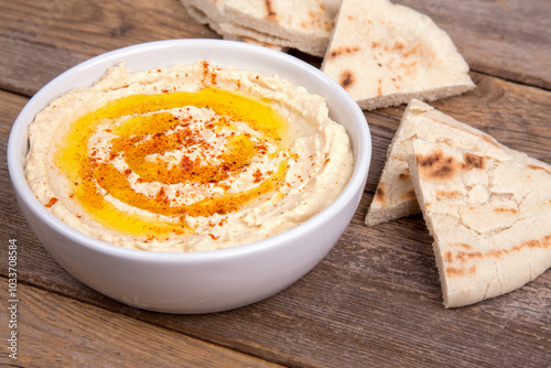 A bowl of creamy hummus drizzled with olive oil and sprinkled with paprika, accompanied by slices of warm pita bread arranged on a rustic wood surface.