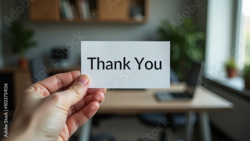 A person holding a thank you note in an office setting with plants, a desk, and a laptop visible during daylight hours
