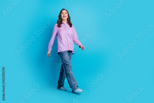 Full length photo of charming positive woman wear violet shirt walking empty space isolated blue color background