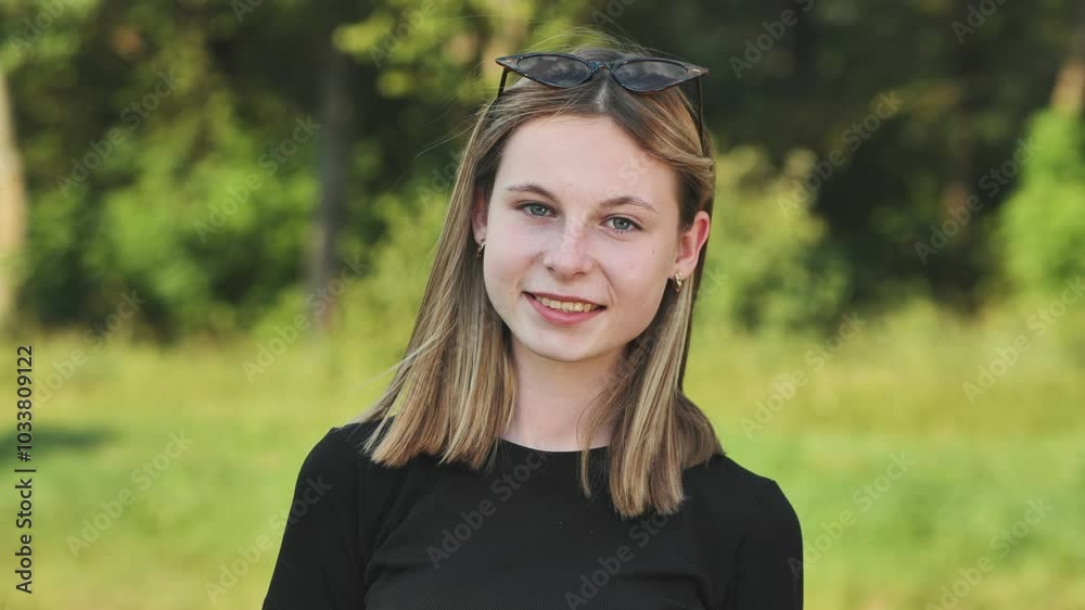 Teenage girl with blonde hair and sunglasses is smiling outdoors with a green background. Teenage girl with blonde hair and sunglasses is smiling outdoors with a green background.