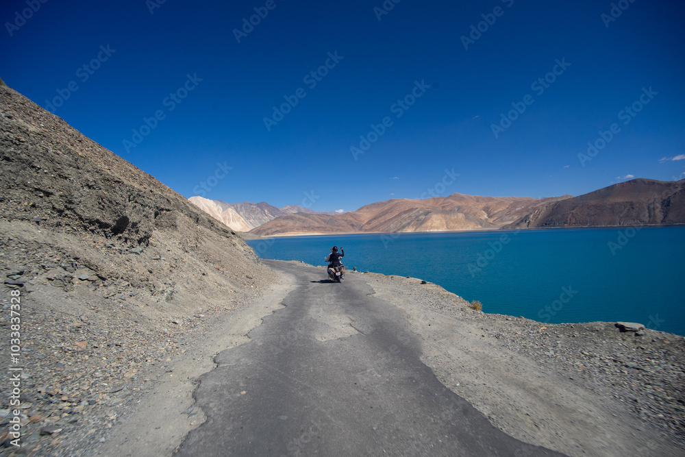 Motorcyclist riding on a scenic mountain road beside a vivid blue lake Motorcyclist riding on a scenic mountain road beside a vivid blue lake