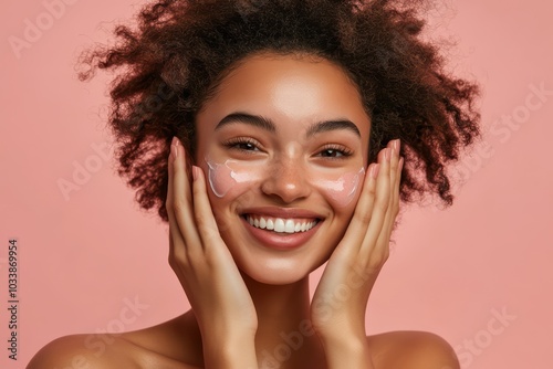 A joyful young woman applying skincare product with a radiant smile against a pastel pink backdrop, showcasing beauty and self-care.