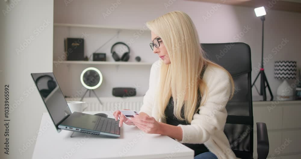 Young beautiful white blonde woman in casual clothes and glasses shopping online sitting at laptop and entering data holding credit card in hands Young beautiful white blonde woman in casual clothes and glasses shopping online sitting at laptop and entering data holding credit card in hands