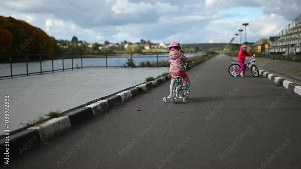 Two little girls riding their bikes on a paved path next to a lake. One girl wears a pink jacket and the other a light purple jacket. The girls are wearing helmets.