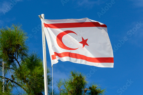 The northern Cyprus flag waving proudly against a clear blue sky with green pine trees in the background on a sunny day