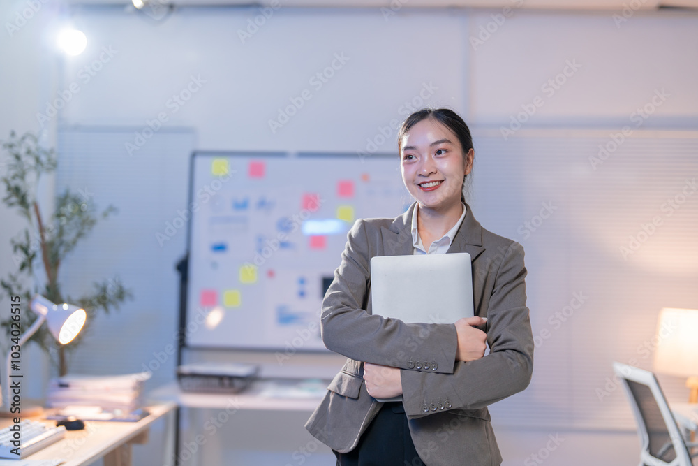 Businesswoman is smiling while holding a laptop in an office at night. She is standing in front of a whiteboard with sticky notes Businesswoman is smiling while holding a laptop in an office at night. She is standing in front of a whiteboard with sticky notes