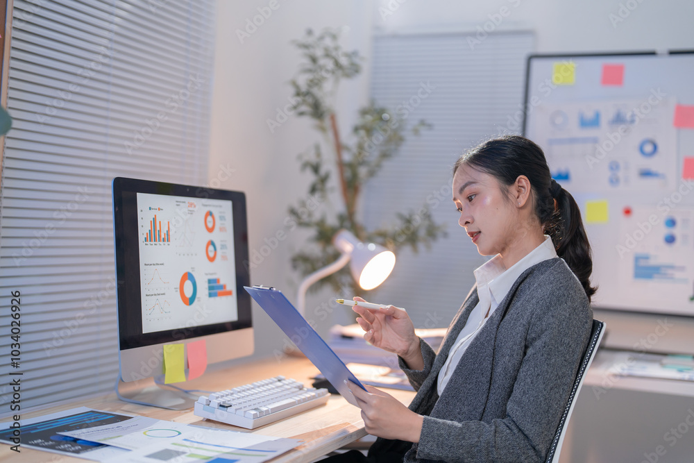 Businesswoman is analyzing charts and graphs on her computer and notepad, working late in the office to prepare a presentation Businesswoman is analyzing charts and graphs on her computer and notepad, working late in the office to prepare a presentation