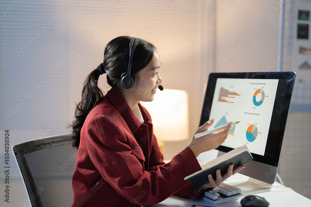 Businesswoman is working late using a headset and computer while holding a notebook and pen. She is pointing at charts on the screen Businesswoman is working late using a headset and computer while holding a notebook and pen. She is pointing at charts on the screen