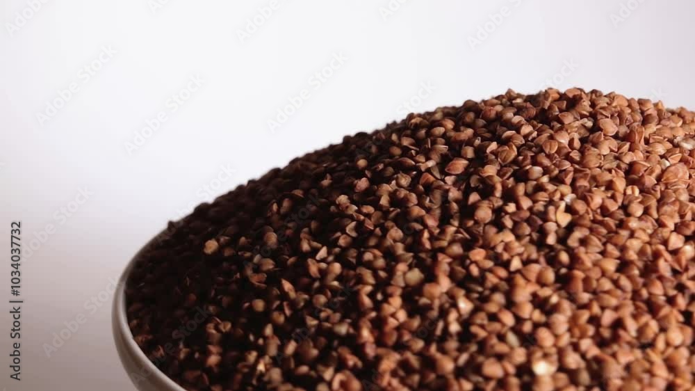 Dry Uncooked Brown Buckwheat Groats Heap on White Plate Rotating against White Background. Raw Large Buckwheat Grains. Russian, Ukrainian and Belarusian Culture Dry Uncooked Brown Buckwheat Groats Heap on White Plate Rotating against White Background. Raw Large Buckwheat Grains. Russian, Ukrainian and Belarusian Culture