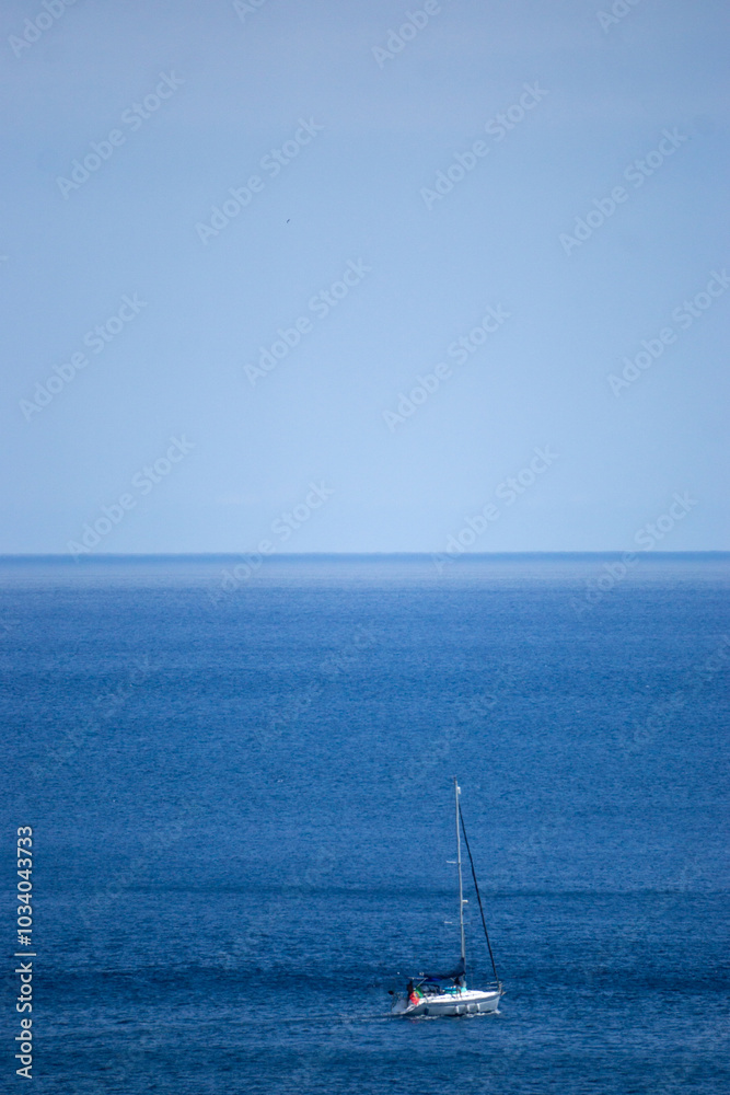A small sailboat with a Portuguese flag floats on the calm waters of the Atlantic Ocean, taken off the coast of the Azores islands. The vast, clear blue sea stretches into the horizon. A small sailboat with a Portuguese flag floats on the calm waters of the Atlantic Ocean, taken off the coast of the Azores islands. The vast, clear blue sea stretches into the horizon.