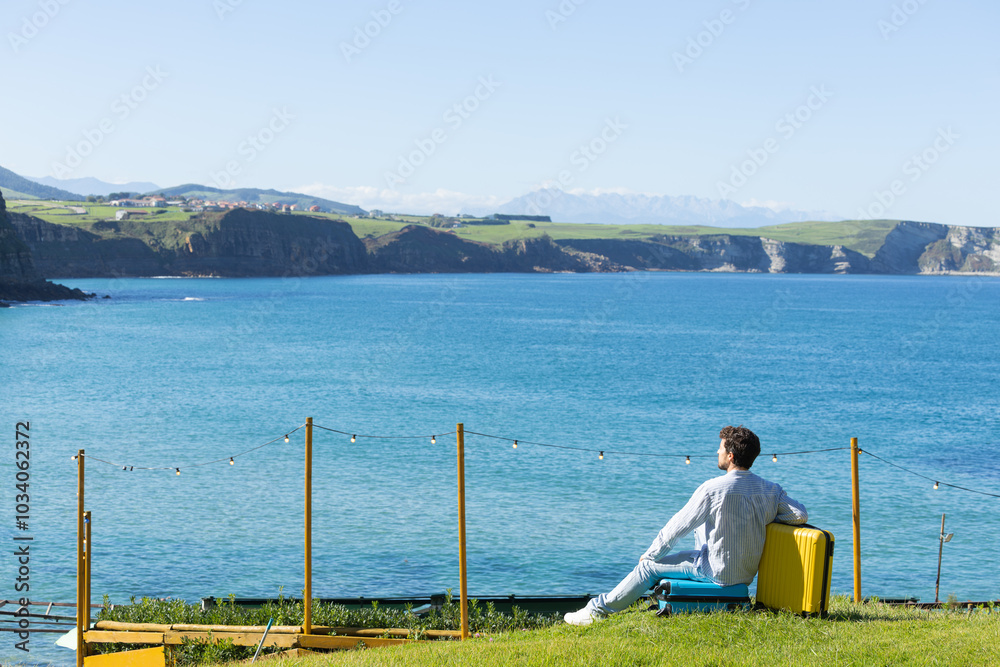 Fototapeta premium Single young adult man resting leaning on his yellow travel suitcase with the background of the sky and the ocean or sea, rest vacation, traveling the world, traveling alone 