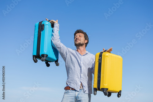 Happy and euphoric single young adult man with his yellow and blue travel suitcases with the background of blue sky and ocean or sea, resting vacation, traveling the world, traveling alone, having fun