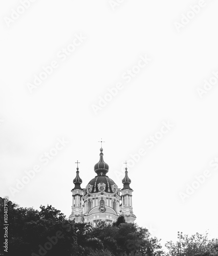 A black-and-white image of St. Andrew's Church in Kyiv, Ukraine, with ornate domes rising above trees, set against a bright, overcast sky, highlighting its baroque architecture.
 photo