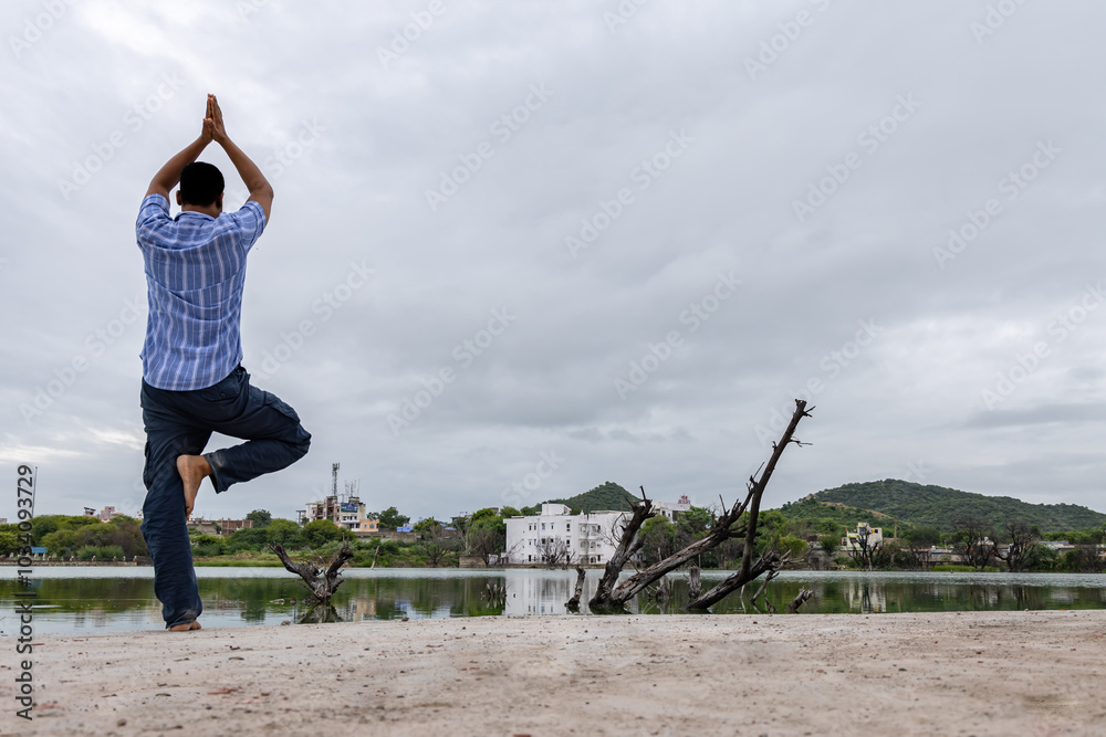 young man meditating and performing yoga near pristine calm lake with dramatic sky at morning young man meditating and performing yoga near pristine calm lake with dramatic sky at morning