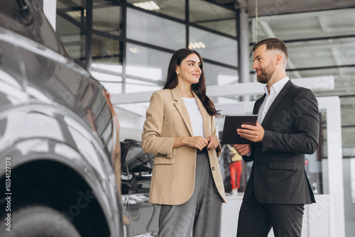 Young woman customer female buyer client wears beige jacket consult with salesman hold tablet choosing auto want to buy new automobile in car showroom vehicle salon dealership store motor show.