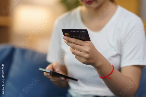 Woman is holding a credit card and using a smartphone for online shopping on a comfortable sofa at home